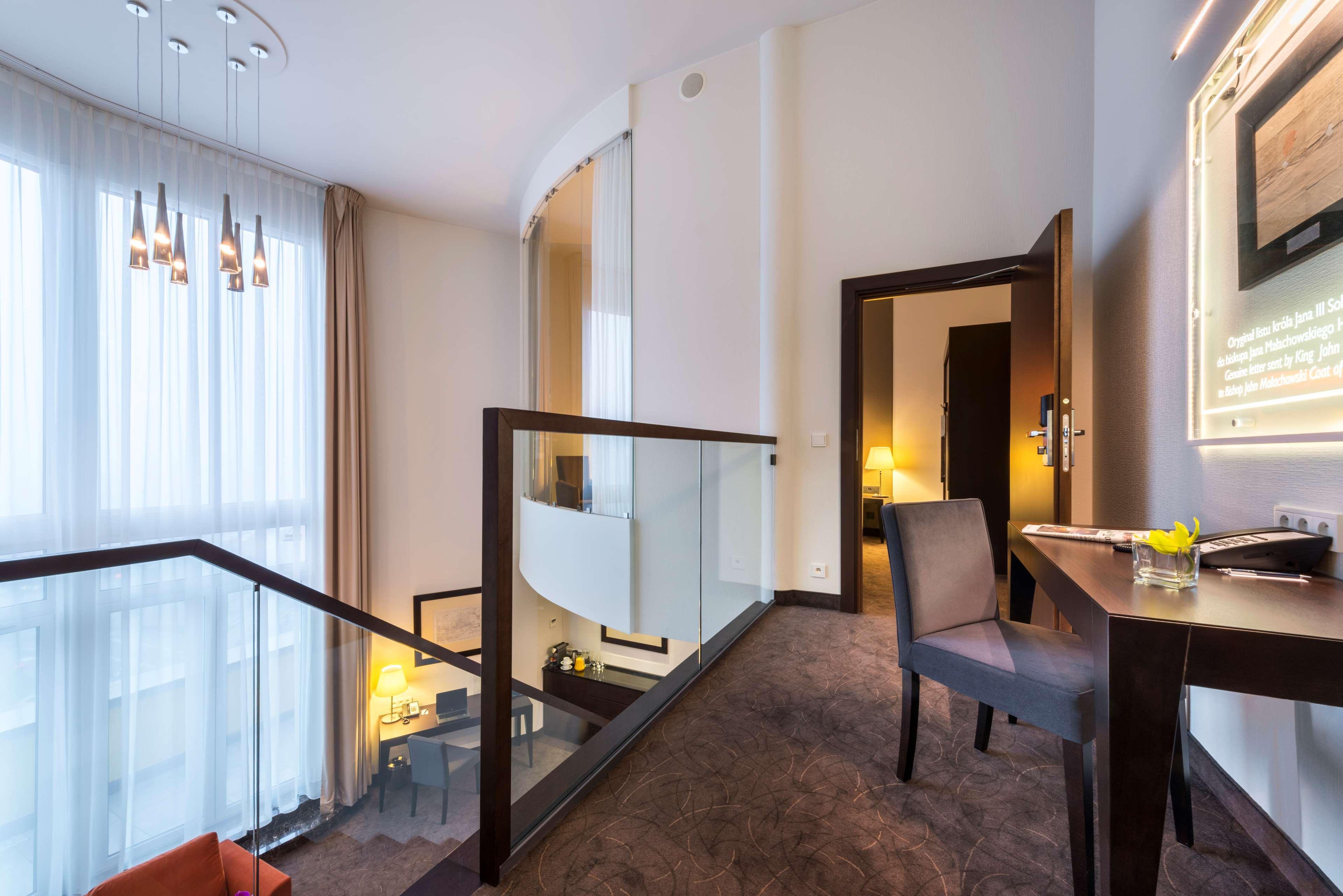 Radisson Blu Sobieski Varsavia Esterno foto The photo depicts a modern interior space, likely a hotel suite or a stylish apartment. There is a staircase leading to an upper level, with a glass railing providing an open atmosphere. The lighting is bright, coming from large windows adorned with 