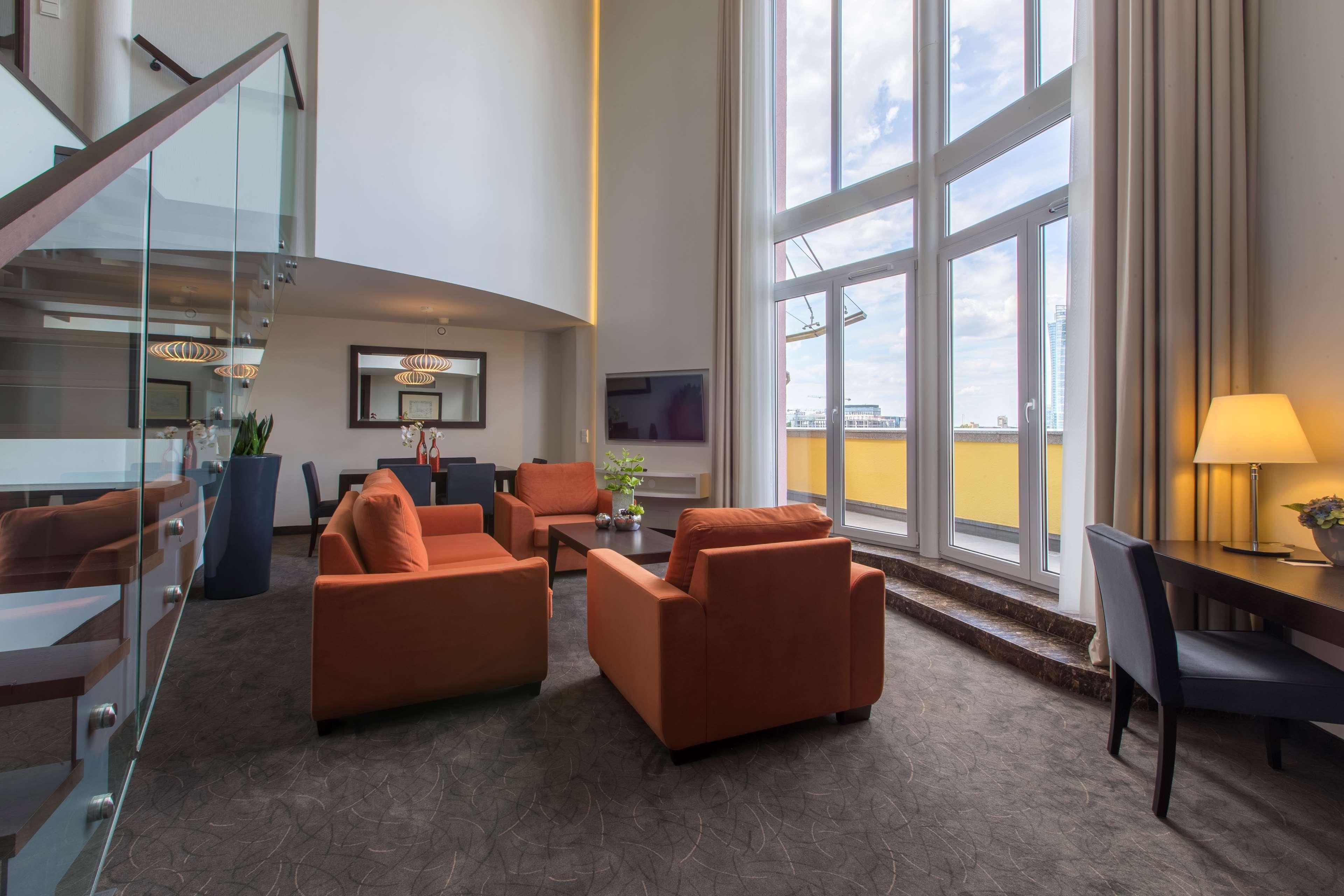 Radisson Blu Sobieski Varsavia Esterno foto The photo shows an elegant and spacious living area. There are four orange chairs arranged around a coffee table, creating a cozy seating space. A large window with multiple panes allows natural light to flood the room, and there are tall curtains th
