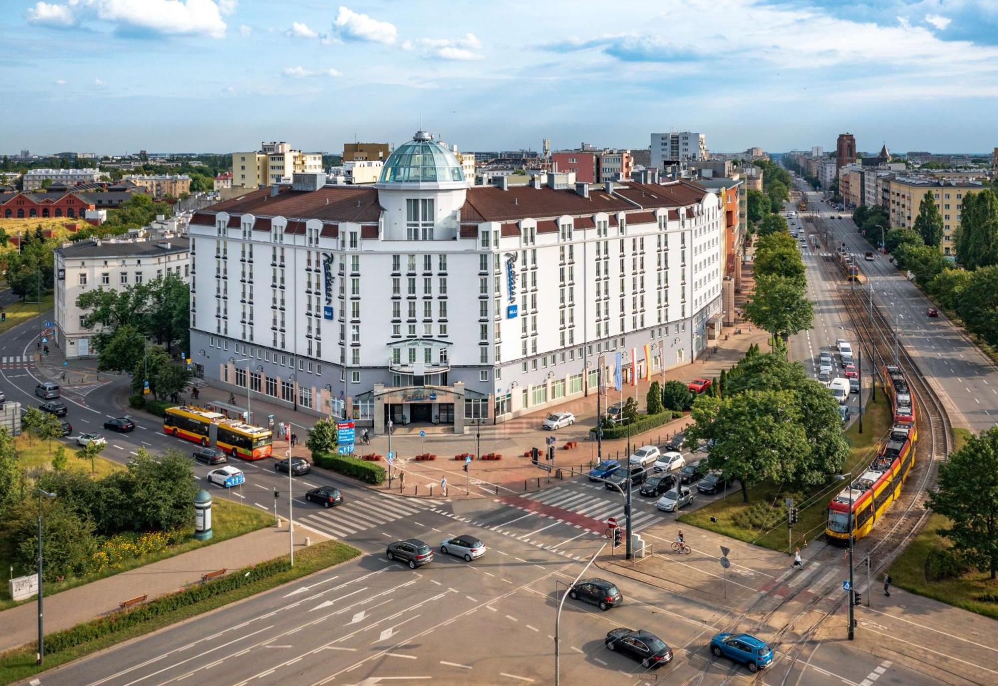 Radisson Blu Sobieski Varsavia Esterno foto The photo shows a large, modern building with a distinctive architectural design, featuring a rounded dome on top. It's situated at a crossroads, surrounded by greenery and several roads. In front of the building, there are a few trams visible, indic